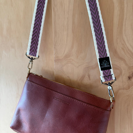 Brown leather clutch with purple and white strap on a wooden surface