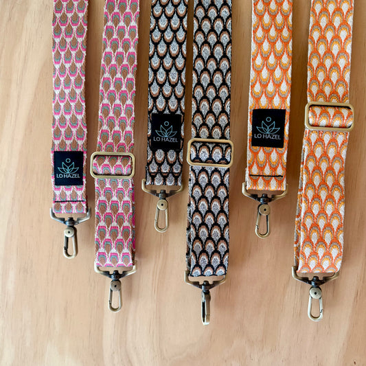 Four patterned bag straps with metal buckles on a wooden surface, featuring the brand 'LO HAZEL'.